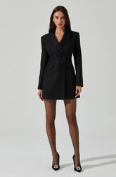 A woman is wearing a black double-breasted blazer-style coat with wide lapels and long sleeves. The coat is tailored and reaches mid-thigh length. She is also wearing sheer black tights and black high heels with decorative embellishments on the toes.